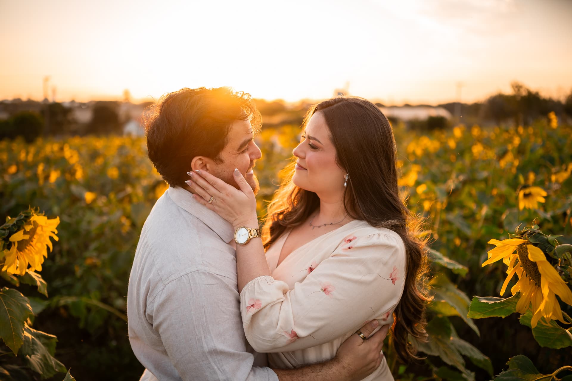 Monique e Thiago abraçados em um campo de girassóis ao pôr do sol