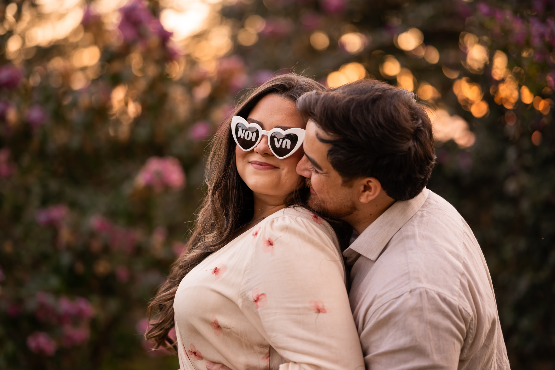 Monique com óculos de noiva e Thiago a abraçando entre flores